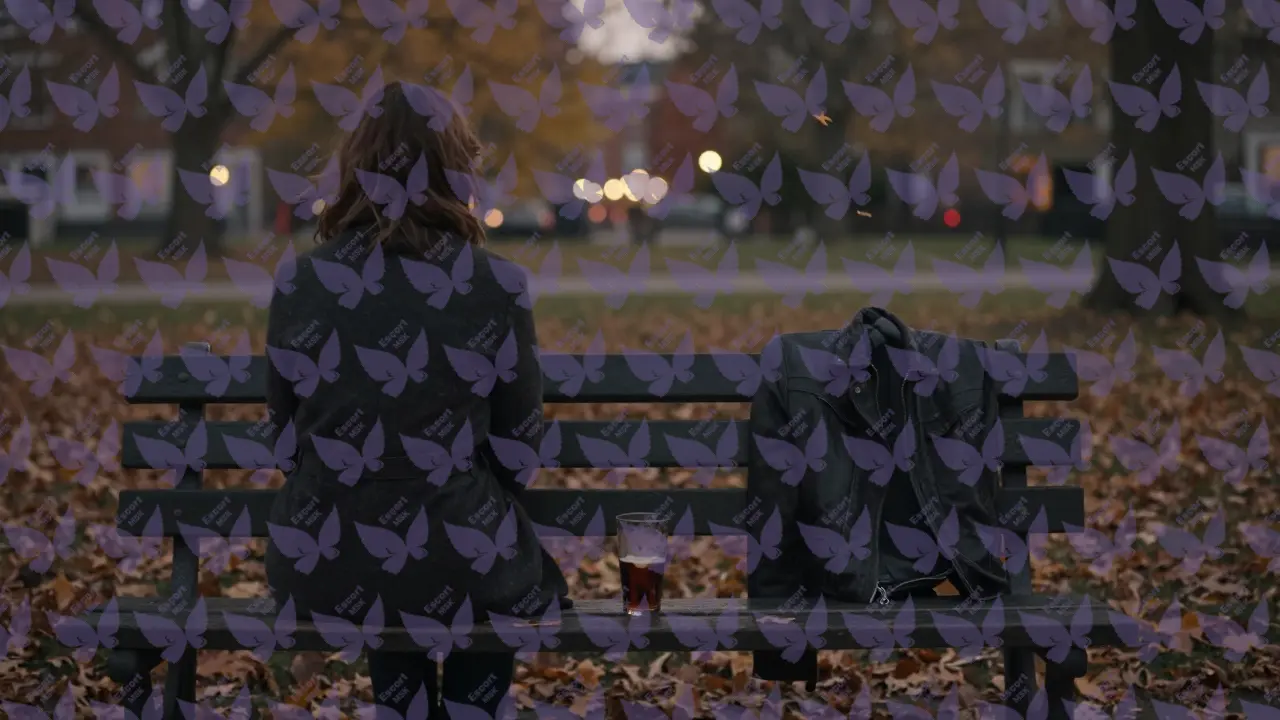 Пустая скамейка в парке с пальто на спинке, вечер, листья падают, городские огни вдалеке.